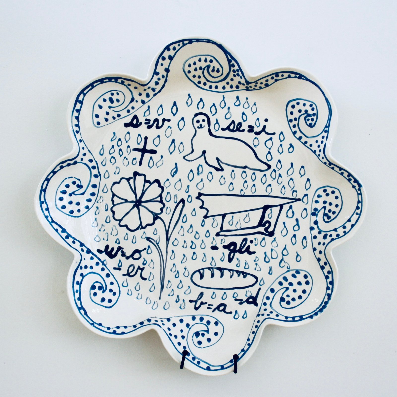 How to Read: Fig 4. Pluvial Flooding, 2024   Fibre clay, low-fired glazes 46 x 46 x 7cm  (18 x 18 x 2.7 inches)  Reading: What we see: a back ground of rain drops framed by undulating waves. What we are being asked to decipher – line drawings and letters. Reading left to right, top to bottom. A plus sign, but we are also told (s=v) thus plus becomes pluv; next an image of seal, here (se=i), thus ial.  Our first word? PLUVIAL; next line, a flower, but, w=o and remove - er (floo) then a silhouette of gliding (–gli) thus ding, FLOODING; Finally, a loaf of bread, however, if  b=a and we are removing -d we have AREA. We are being warned that this is a Pluvial Flooding Area!